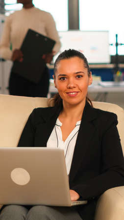 Hispanic business woman smiling at camera sitting on couch typing on computer while diverse...