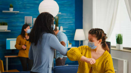 Happy multiracial friends with protective mask touching elbow keeping social distancing greating...