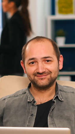 Manager sitting on couch in front of camera smiling while multiethnic team working in background with financial reports. Diverse coworkers talking about startup financial company in modern office.の写真素材