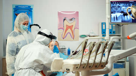 Pediatric dentist in protective suit lighting the lamp until examination while little patient opening mouth. Medical team talking wearing face shield, coverall suit, mask and gloves during coronavirusの写真素材