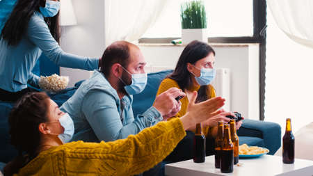 Multiracial people celebrate winning video game in home living room with joystick wearing face mask keeping social distancing in time of corona outbreak. Diverse friends enoying beer and chips.の写真素材
