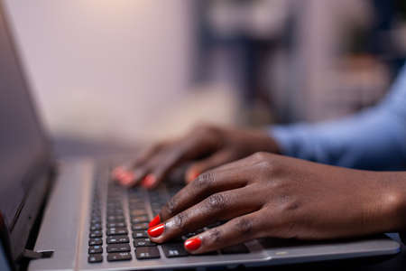 Side view of dark skinned business woman typing on laptop sitting at desk late at night. Employee using modern technology network wireless doing overtime.の写真素材