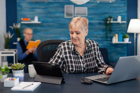 Senior business owner working from home office using portable computer. Elderly woman in home living room using moder technoloy laptop tablet pc for communication sitting at desk indoors.の写真素材
