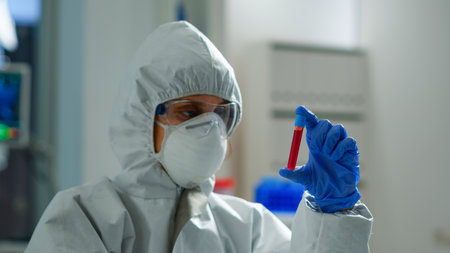 Biochemist in coverall holding blood sample analysing chemical reaction working in medical lab. Scientist examining vaccine evolution using high tech for treatment development against covid19 virusの写真素材