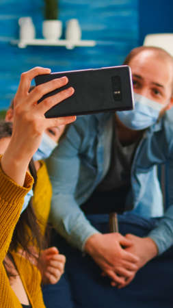 Happy friends from diverse cultures and races taking selfie wearing protection masks during new normal party having fun together keeping social distance, spending free time in living roomの写真素材