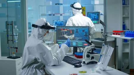 Chemist doctor in ppe suit working at pc while lab technician bringing her blood samples. Team of...