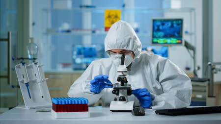 Scientist in ppe suit making adjustments and looking through laboratory microscope, typing on pc....