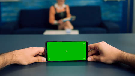 Pov of man hands holding in horizontal mode smartphone with mock up green screen chroma key display standing on desk in office studio. Freelancer using isolated phone for browsing social networksの写真素材