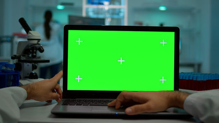 Pov shot of microbiologist typing on laptop with green chroma key display sitting at desk working reading virus symptoms. In background lab researcher analysing vaccine developent examining samplesの写真素材