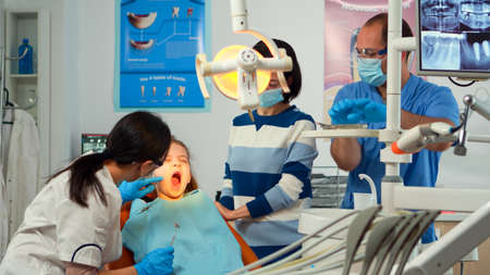 Pedicatric dentist doctor working in dental unit with nurse and little girl patient. Stomatologist speaking to mother of girl with toothache sitting on stomatological chair while man preparing tools.の写真素材