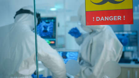 Chemists in coverall trying to develop vaccine using tablet standing behind the glass wall working in danger area of lab. Scientists examining virus evolution researching for treatment against covid19の写真素材