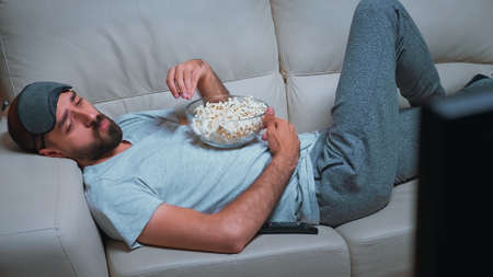 Tired man relaxing on sofa in front of television eating popcorn while watching movie show....