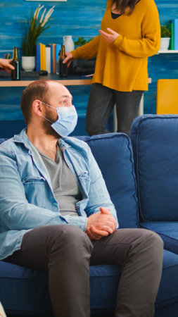 Diverse friends discussing during new normal party wearing protection mask, socializing keeping distance sitting on sofa drinking beer and eating snaks. Group of multi ethnic people enjoying free timeの写真素材