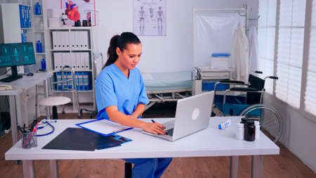 Therapist writing list of consulted patients online, making research, noting, taking useful information on clipboard. Doctor assistant in uniform working on laptop in modern hospital office.の写真素材
