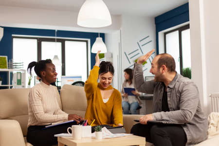 Diverse team of successful young entrepreneurs sharing high-five celebrating success sitting on couch in start up office room. Multiethnic business team with laptop and papers excited about projectの写真素材