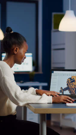 African engineer working late at night on a 3D model of industrial gears at laptop doing overtime in...