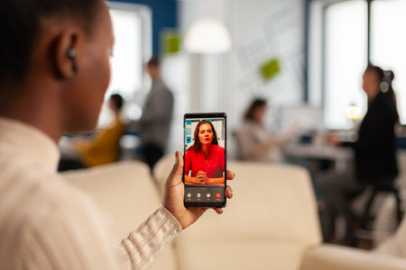 Over the sholder of black employee discussing with partner on video call using phone in start up...