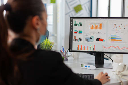 Businesswman analysing financial statistics in new normal offfice, wearing face mask as safety...