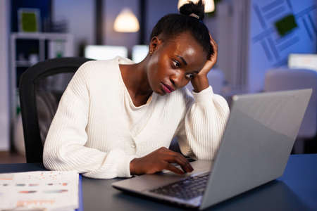 Tired african woman freelancer sitting at desk table analysing economic statistics. Exhausted businesswoman planning financial accounting working in startup business company officeの写真素材