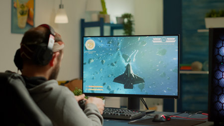 Pro man gamer playing video game at powerful personal computer late night wearing professional headphones. Excited player using wireless controller for virtual tournament gaming space shooter at homeの写真素材