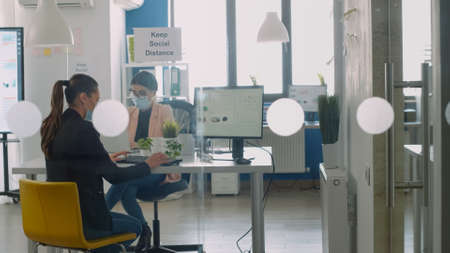 Businesswoman wearing protective face mask working on computer and discussing wiht collegue avout business job. Coworkers keeping social distancing to avoid virus disease during global epidemicの写真素材
