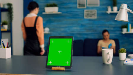 Home studio equipped with tablet computer with mock up green screen chroma key display standing in vertical mode on office desk. In background two collegues talking about internet advertisingの写真素材