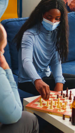 Multiethnic friends playing chess in home living room talking keeping social distancing to prevent illness with covid19 during global pandemic wearing face mask. Diverse people enjoying board gamesの写真素材
