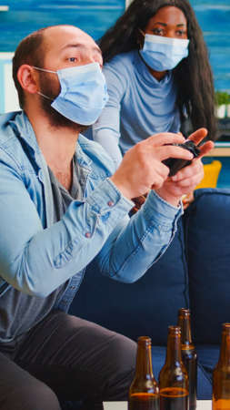 Multiethnic friends celebrating victory playing video games in living room raising hands wearing face mask and keeping social distancing during global pandemic. People enjoying time togetherの写真素材