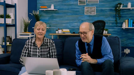 Elderly age couple sitting on sofa waving at camera during videocall with nephews using laptop in...