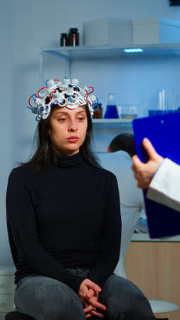 Medic researcher pointing at clipboard explaning treatment against brain disease to patient with eeg headset. Woman sitting in neurological scientific laboratory treating nervous system dysfunctionsの写真素材