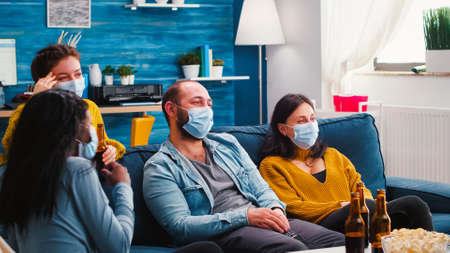 Group of multiracial friends watching comedy show on tv laughing enjoying spending time together wearing face mask to prevent infection with covid 19 during global pandemic having fun sitting on couchの写真素材
