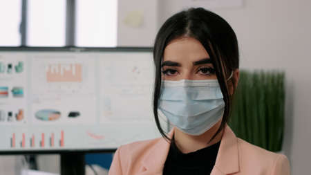 Closeup of businesswoman wearing medical face mask to avoid virus infection during coronavirus lockdown. Entrepreneur looking into camerea while sitting in startup company office.の写真素材