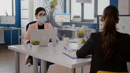 Freelancer talking with coworker about business startegy while sitting in new normal office wearing protective face mask to prevent infection  . Team respecting social distancingの写真素材