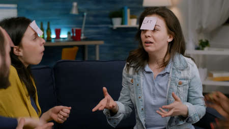 Multiethnic friends sitting on couch late at night playing fuuny guess who game using sticky notes.の写真素材