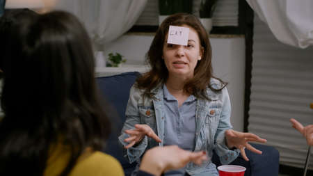 Creative woman playing who i am with her friends with sticky papers attaching to foreheads, enjoying time together. A group of people hanging out in the living room during funny party.の写真素材