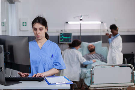Front view of medical asisstant typing illness expertise on computer while in background physician...