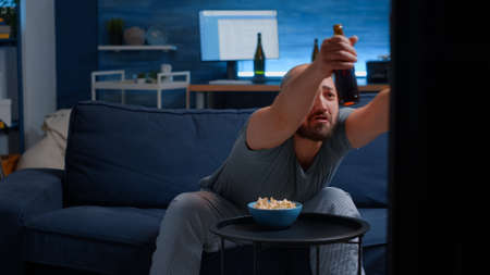 Man holding remote controller eating popcorn watching sports on TV supporting team raising hands...