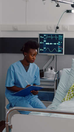 Sick woman resting in bed while afro-american nurse monitoring disease writing medical healthcare treatment on clipboard. Assistant discussing illness therapy working in hospital wardの写真素材