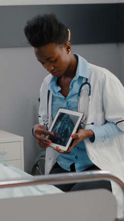 Afro american practitioner doctor analyzing bones radiography to sick man using tablet computer in hospital ward. Patient with nasal oxygen tube resting in bed during healthcare recoveryの写真素材