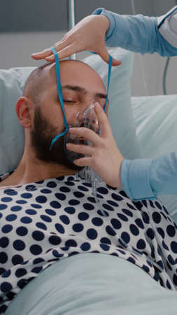 Patient sitting in bed while woman doctor putting oxygen mask monitoring respiratory illness. Physician medic writing sickness treatment working in hospital ward during recovery appointmentの写真素材