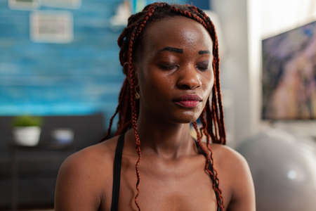 Black woman training body and mind meditating in lotus pose keeping eyes closed, sitting on yoga mat in home living room for calm, healthy harmony lifestyle dressed in sportwear.の写真素材