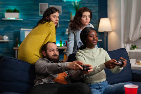 Multi ethnic friends feeling good during oline games challange using wireless controller. Mixed race group of people hanging out together having fun late at night in living room.の写真素材
