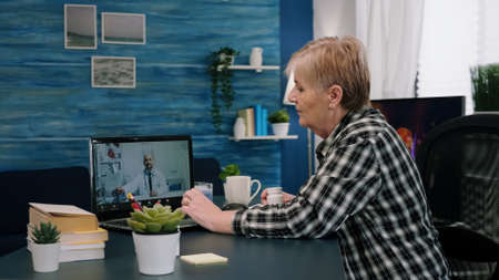 Remote young physician using webcam prescribing medicine to sick aged woman sitting in living room via internet. Patient talks with therapist by videoconference call application, holding pills flaskの写真素材