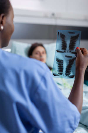 Close-up of afro american nurse analyzing lungs x-ray checking healdcare recovery. Sick woman patient resting in bed discussing disease treatment in hospital ward during therapy consultationの写真素材