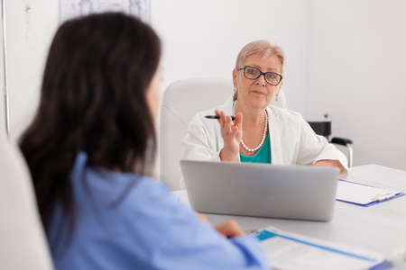 Portrait of senior physician woman discussing pills medication treatment with medical nurse sitting in hospital conference meeting room. Doctor in white coat writing sickness diagnosis on clipboardの写真素材