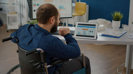 Disabled handicapped immobilized paralyzed manager man using computer and tablet in same time working in business start-up office sitting in wheelchair. Busy multitasking employee typing searchingの写真素材