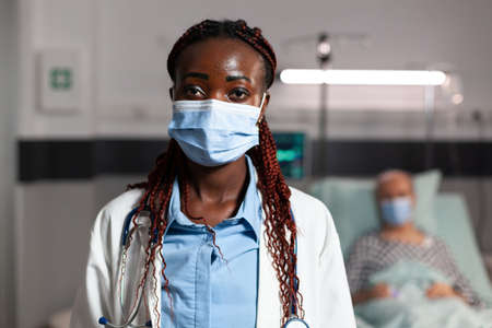 African american doctor health care specialist wearing chirurgial mask in hospital room with sick patient btreathing with help from oxygen tube and iv drip attached.の写真素材
