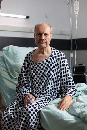 Portrait of sad, unwell senior man sitting on the edge of hospital bed with iv drip attached and breathing with help from oxygen mask, looking at camera.の写真素材