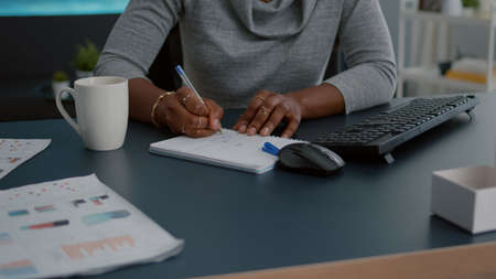 Closeup of student with black skin writing communication homework on notebook while sitting at desk in living room. Young woman studying math on elearning platform doing homework during high schoolの写真素材