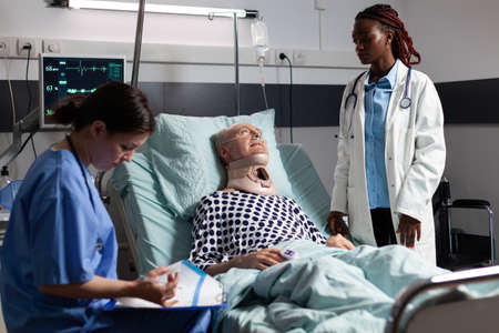 Injured senior man with neck brace laying in bed suffering after accident, discussing with african...